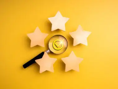 Magnifying glass over a checkmark symbol surrounded by five wooden stars on a yellow background.