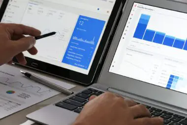 Hands working on a laptop and tablet displaying Google Analytics dashboards and data charts.
