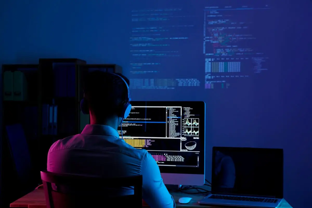 A programmer sits at a desk in a dark room with blue lighting, working on code displayed on a computer screen.