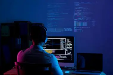 A programmer sits at a desk in a dark room with blue lighting, working on code displayed on a computer screen.